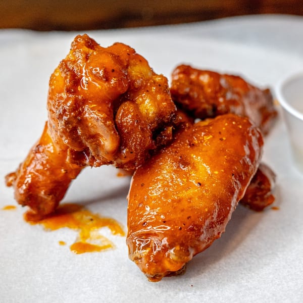 Close-up of three saucy, orange-glazed chicken wings on white parchment paper, with a small cup in the background.