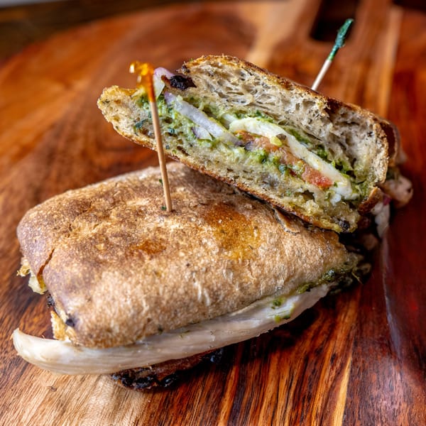 A cut panini sandwich with pesto, cheese, and vegetables, held by two toothpicks on a wooden board.