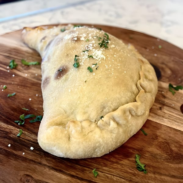 A golden-brown calzone, topped with parmesan and parsley, rests on a wooden board.