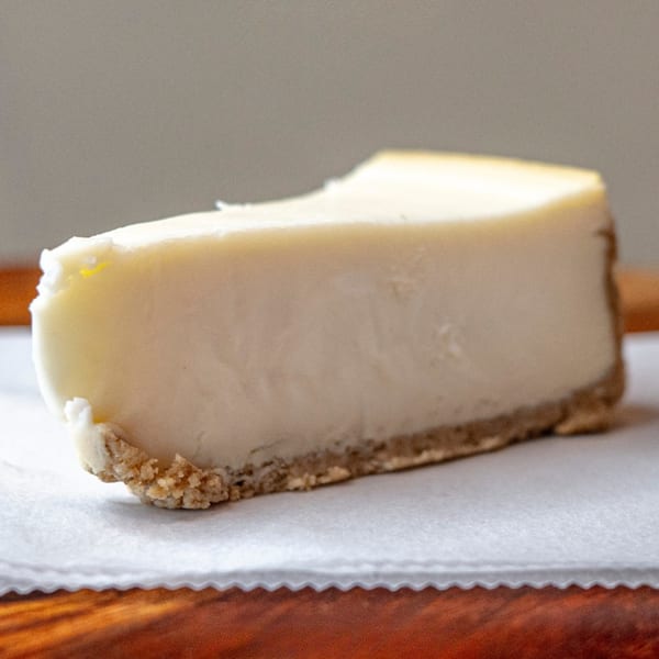 A slice of classic cheesecake with a graham cracker crust on white parchment paper.
