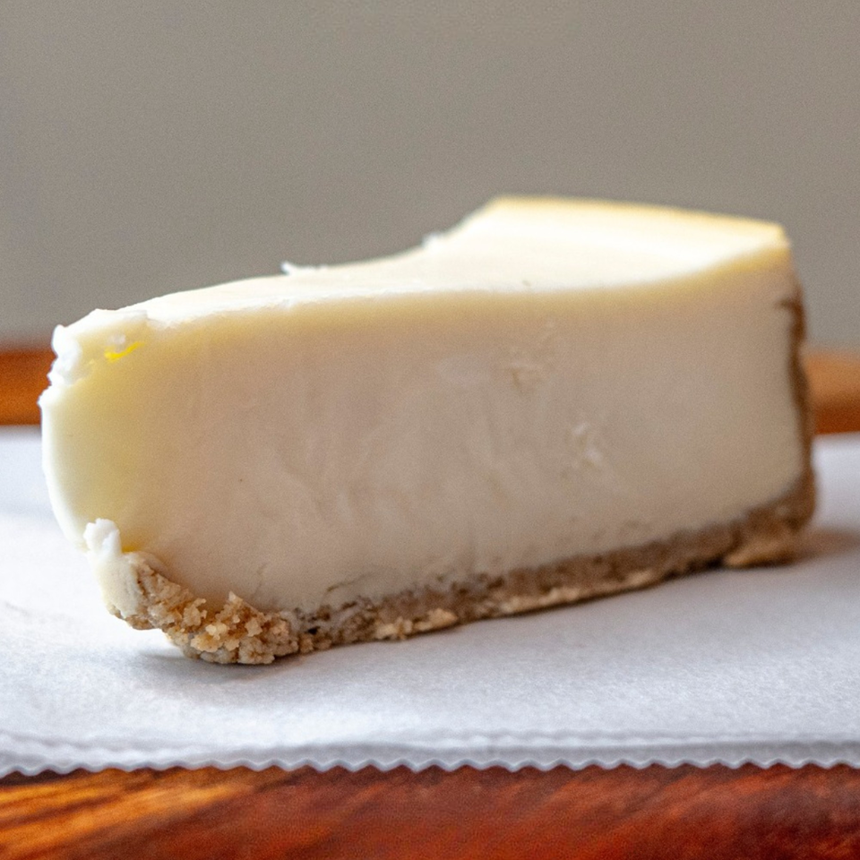 A slice of classic cheesecake with a graham cracker crust on white parchment paper.